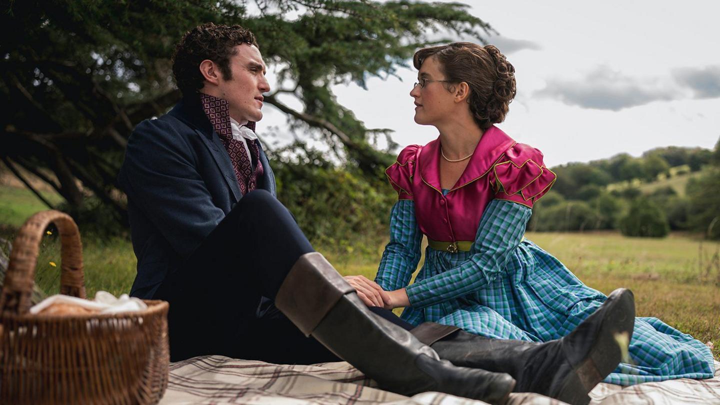 Mary Bennet played by Ella Bruccoleri is photographed alongside Tom Hayward, played by Donal Finn. The fictional couple are sat on a picnic blanket holding hands, staring into each other's eyes. A wicker picnic basket can be seen in the bottom left of the image. Behind them is a large tree.