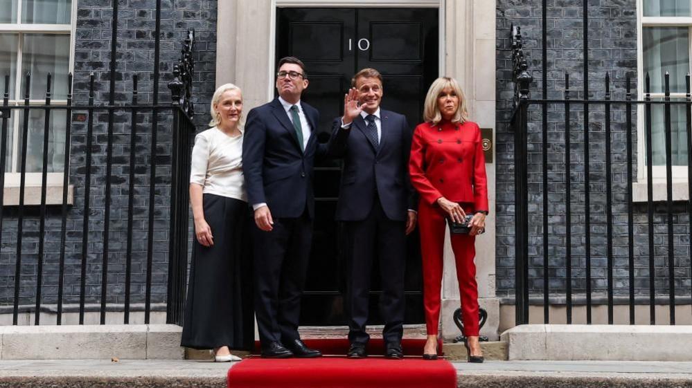 Andy Burnham and Emmanuel Macron stand on a red carpet outside Number 10's black front door, Macron waving and both smiling. Burnham's wife is seen wearing white next to Burnham, while Macron's wife is seen wearing red.