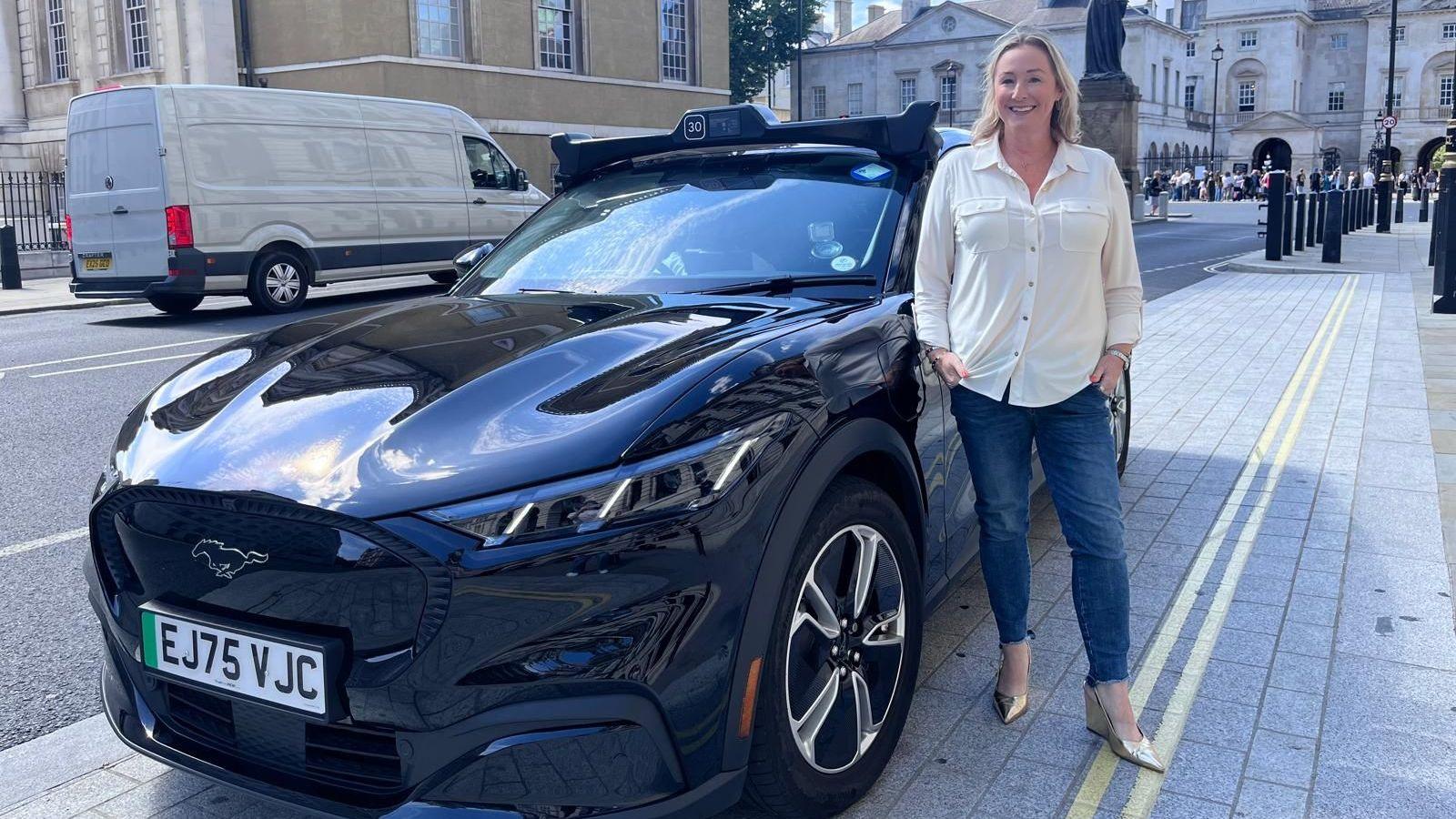 Zoe Kleinman in blue jeans and cream blouse standing next to a dark Wayve self-driving robotaxi on a London pavement.