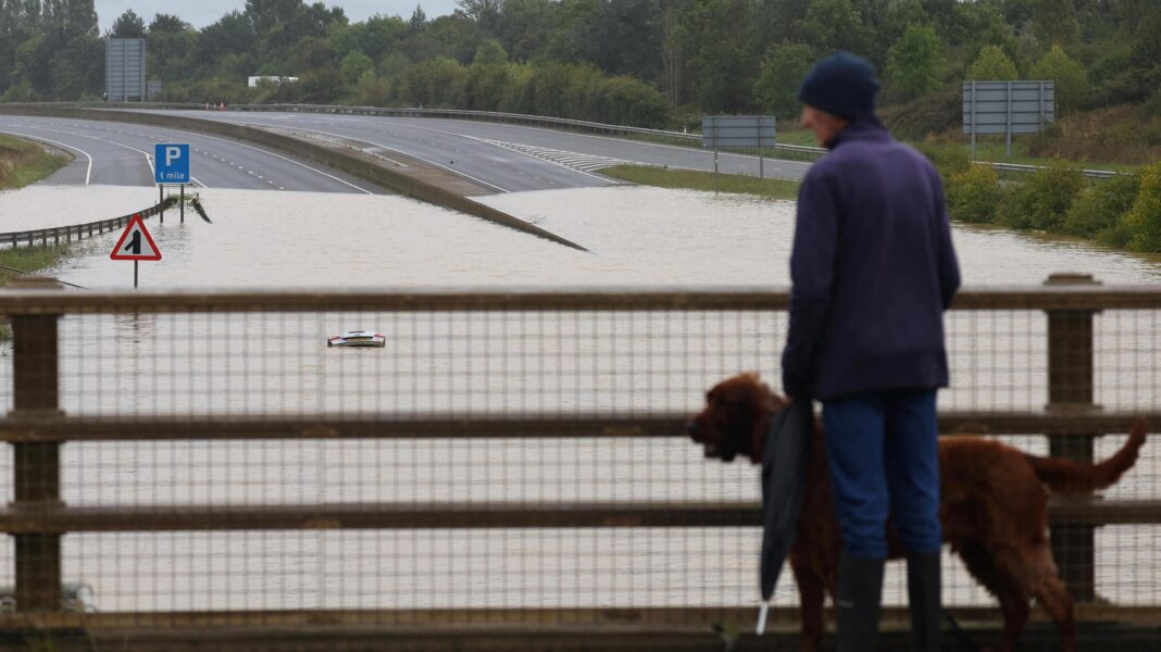 A man and his dog stand on a bridge and look at a flooded road. A car can be seen submerged in the water.