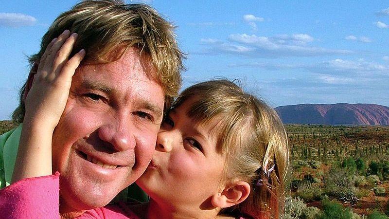 A man with short brown hair and a young girl holding his face and kissing him on his cheek, with Uluru in the background
