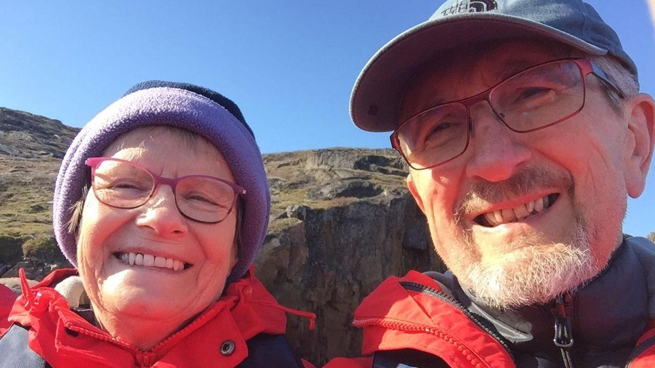Linda and Geoff Kitchen. They are pictured in a selfie outside, in front of a cliff edge. They are both wearing glasses, hats and red waterproof coats and are looking at the camera and smiling.