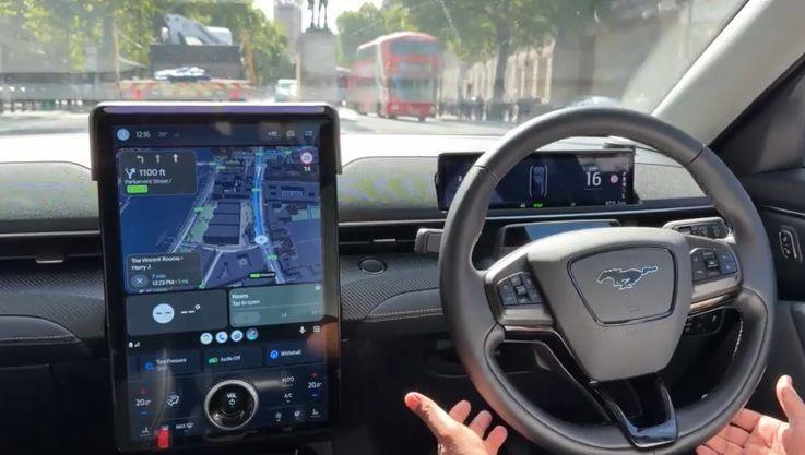 A passenger's view inside a robotaxi as it drives through London, with the hands of the safety driver not touching but around the wheel of the car, and a large black tablet with a map on it showing where the car is going.