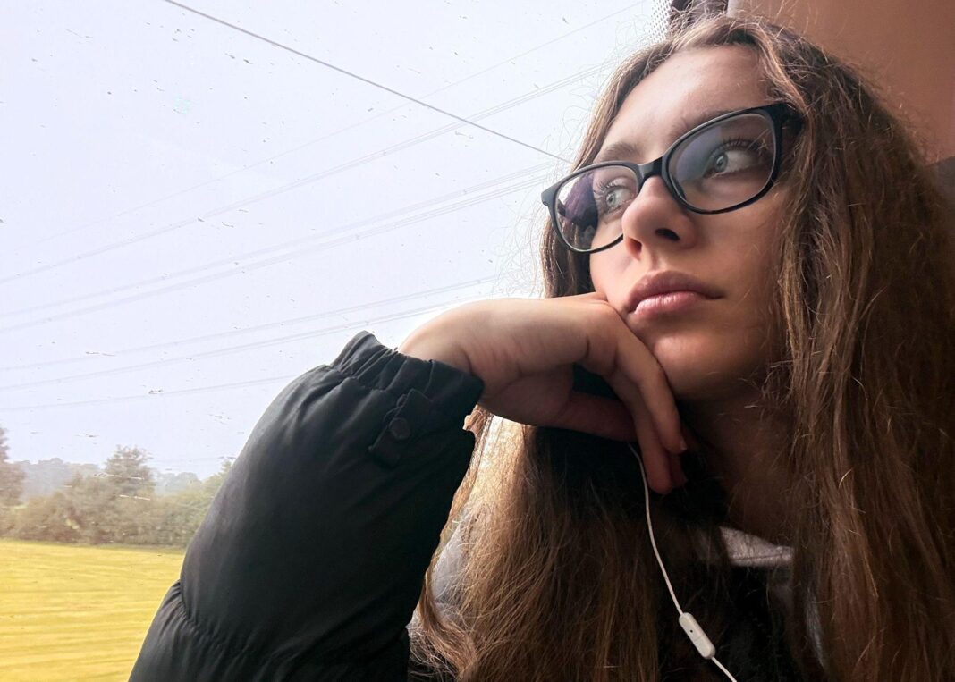 Fresh bedsheets and dusk walks: Expert tips for sleep-deprived freshers A young woman with long brown hair and dark-rimmed glasses stares out of a train window as it moves along. She is wearing a dark jacket and white earphones are visible.