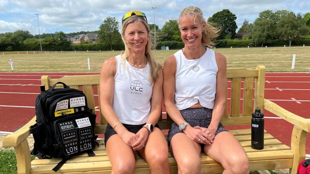 Two women sitting in running outfits on a bench with a running track behind them.