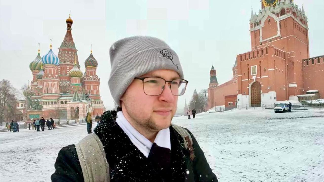 Person in the foreground wearing a grey beanie hat, glasses, a dark coat and a backpack, with snow on clothing and shoulders. Snow-covered cobbled square surrounds the scene. In the background, St Basil’s Cathedral with colourful onion domes stands to the left, and a red brick Kremlin wall with a tower is on the right. Several pedestrians walk across the square, and a parked vehicle is visible near the wall. Snow is falling lightly across the image.
