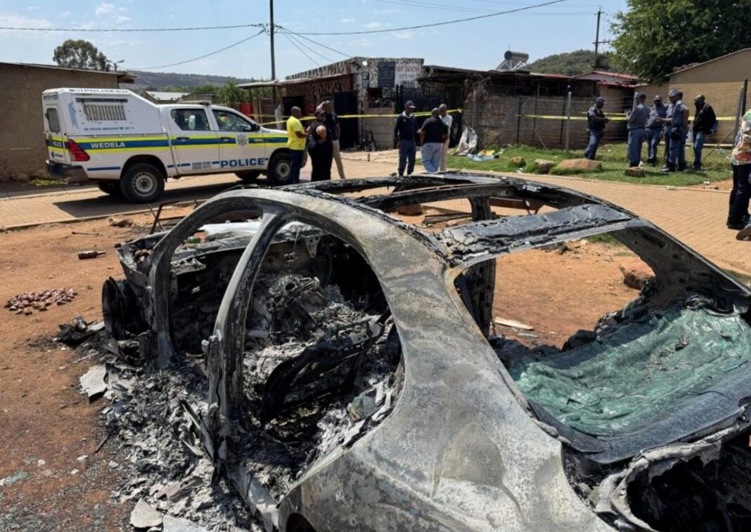 Two mass shootings in South Africa leave 27 dead Shot of a burnt car in Johannesburg, while police officers and stand around it. A police van and a police cordon around a building can be seen in the background.