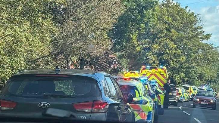 A black car is parked behind six yellow and blue police cars and two yellow and red ambulances on the side of a road
