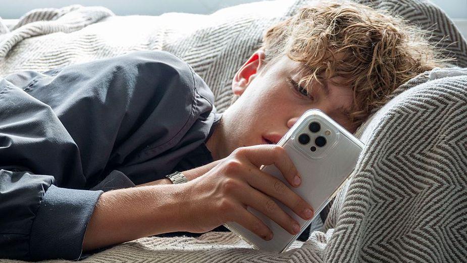 Young person with curly brown hair lying on a sofa, looking at a white phone