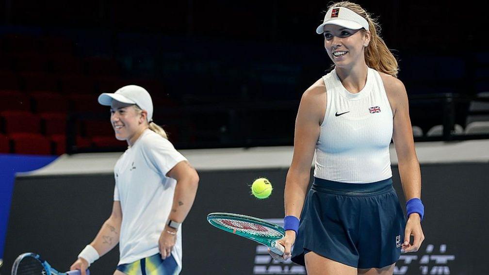 A smiling Sonay Kartal and Katie Boulter in training. Boulter bounces a ball on her racquet.