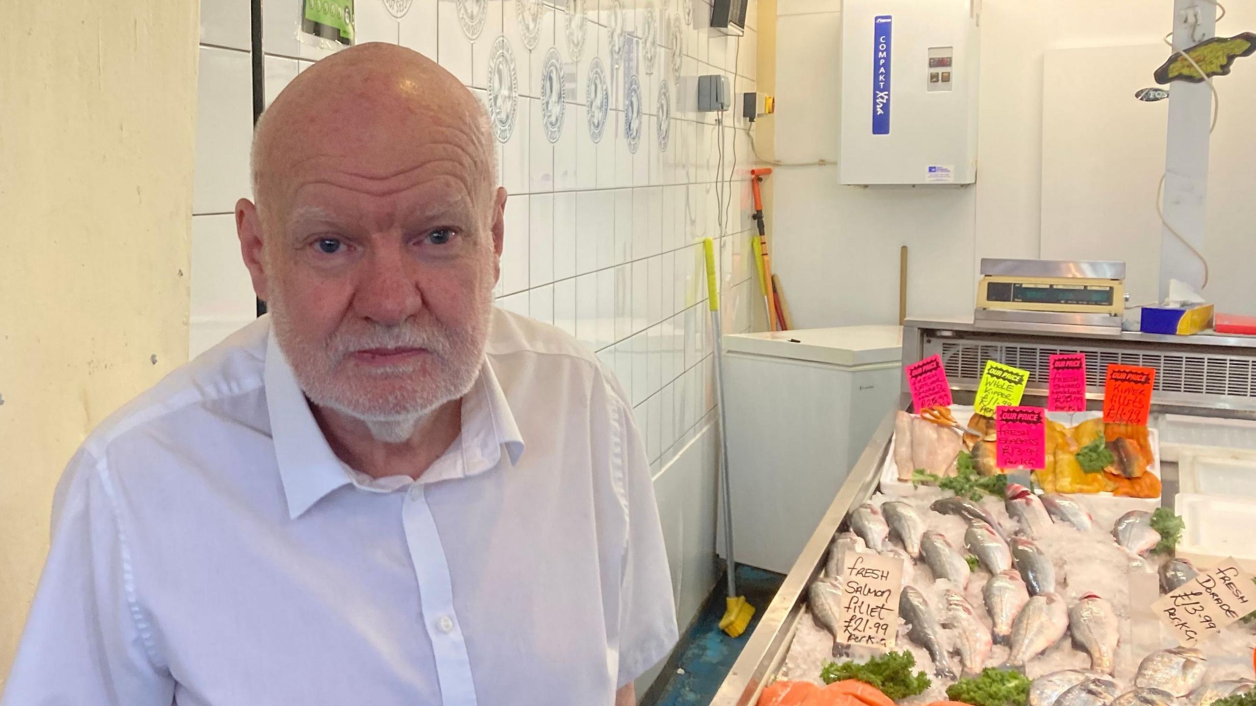 A person stands beside a fish counter inside Coventry Market. The chilled display contains whole fish and fillets arranged on crushed ice, with brightly coloured handwritten price cards showing the names and prices of different products. White tiled walls, weighing scales, refrigeration equipment and cleaning tools are visible behind the counter, creating a traditional fishmonger's shop setting.