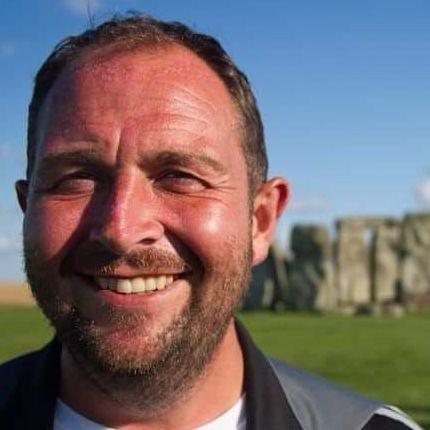 Aaron Howlett smiles in front of stone henge. He has brown short hair and brown beard.