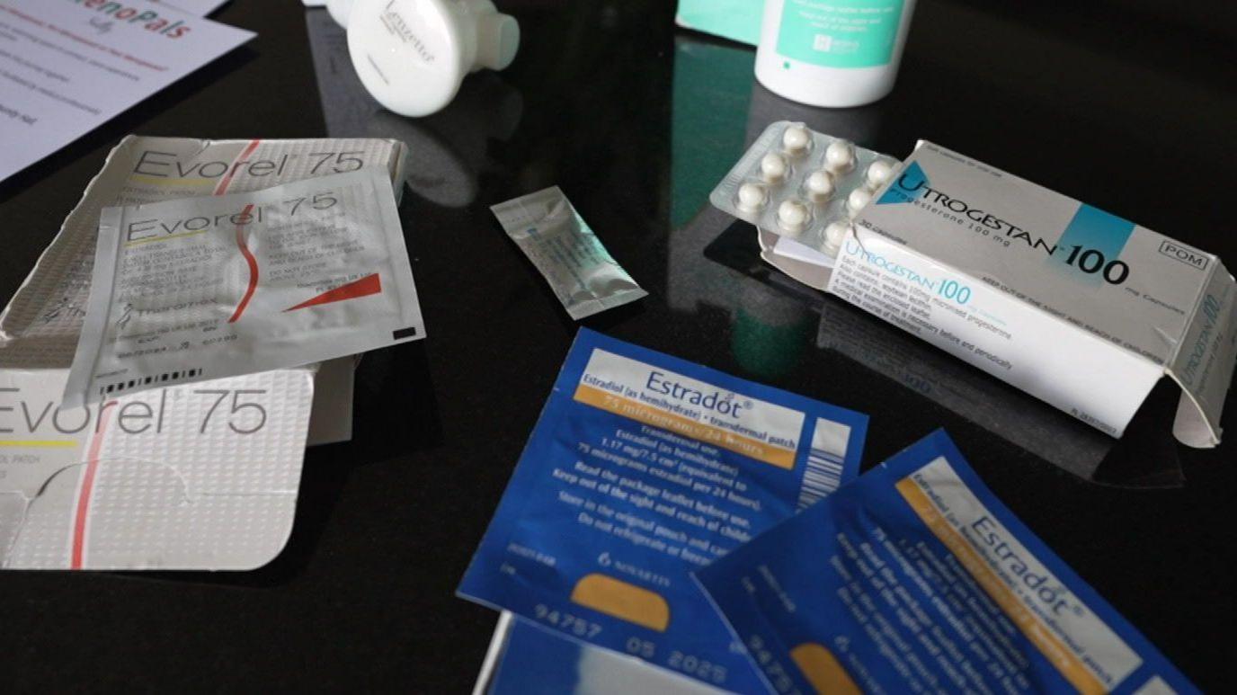 Different types of HRT are laid out on a table, including tablets, patches, spray and gel. In the centre is a small sachet of testosterone gel.