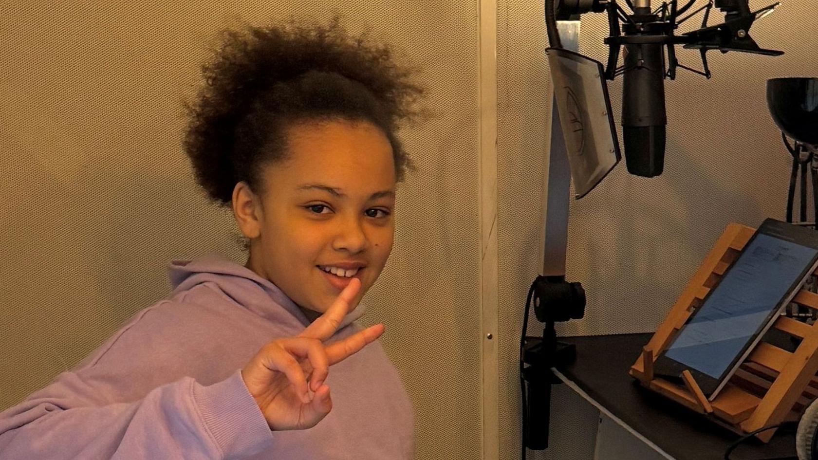 A girl with tied back bushy dark hair, wearing a pink hooded top, smiles as she puts two of her fingers up in a peace sign. She is sitting in a recording booth and a microphone and iPad are set up in front of her.
