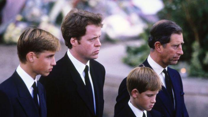 A young Prince William stands next to Earl Spencer, a young Prince Harry and Charles at Diana's funeral. They are all wearing dark suits with ties and are facing ahead solemnly.