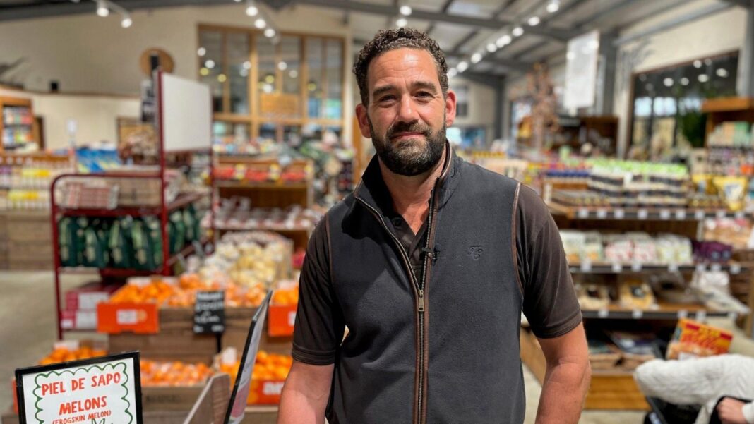 Our shop doors are locked during opening hours after theft Andrew is standing in his farm shop, with shopping aisles behind him. He surrounded by produce on the shelves.