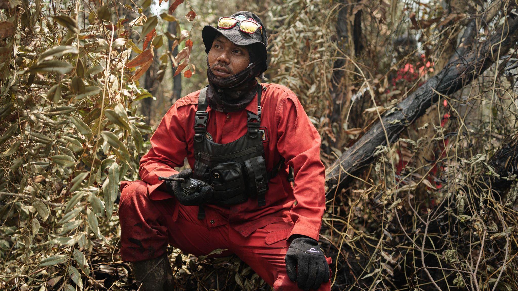 A man wearing red firefighting uniform, tactical vest, gloves, neck gaiter and hat crouches in a forest, holding a handheld radio device
