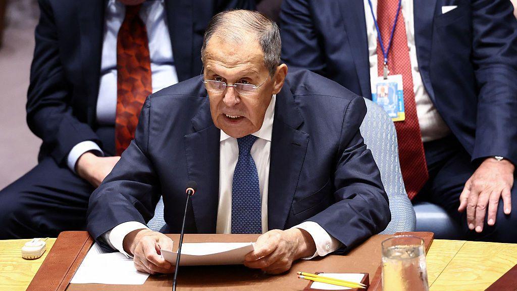 Sergey Lavrov wearing a navy suit, white shirt, and blue tie. He is sat behind a microphone with other diplomats in suits behind him. Lavrov is holding several documents in his hands, wears glasses, and has short grey hair.