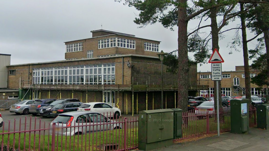 Exterior view of a large, multi-storey brick building beside a road, with tall pine trees lining the pavement. The building has long rows of white-framed windows and a flat roof, with hills visible in the distance beneath an overcast grey sky. The car park behind a red metal fence is full of cars.