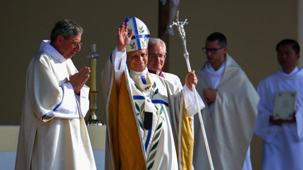 The pope waves to crowds. He is wearing traditional dress including a mitre with a blue cross and leaves on it. He has matching robes which are yellow beneath his arms. His is holding a metal pole with a sculpture of Jesus on the cross at the top. Several people wearing white robes can be seen around the pope