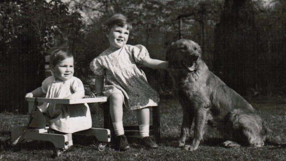 Esther Rantzen as a child (centre) in a black and white photo.  She looks around three years old and is flanked by a younger sibling and a pet dog (which she is stroking)