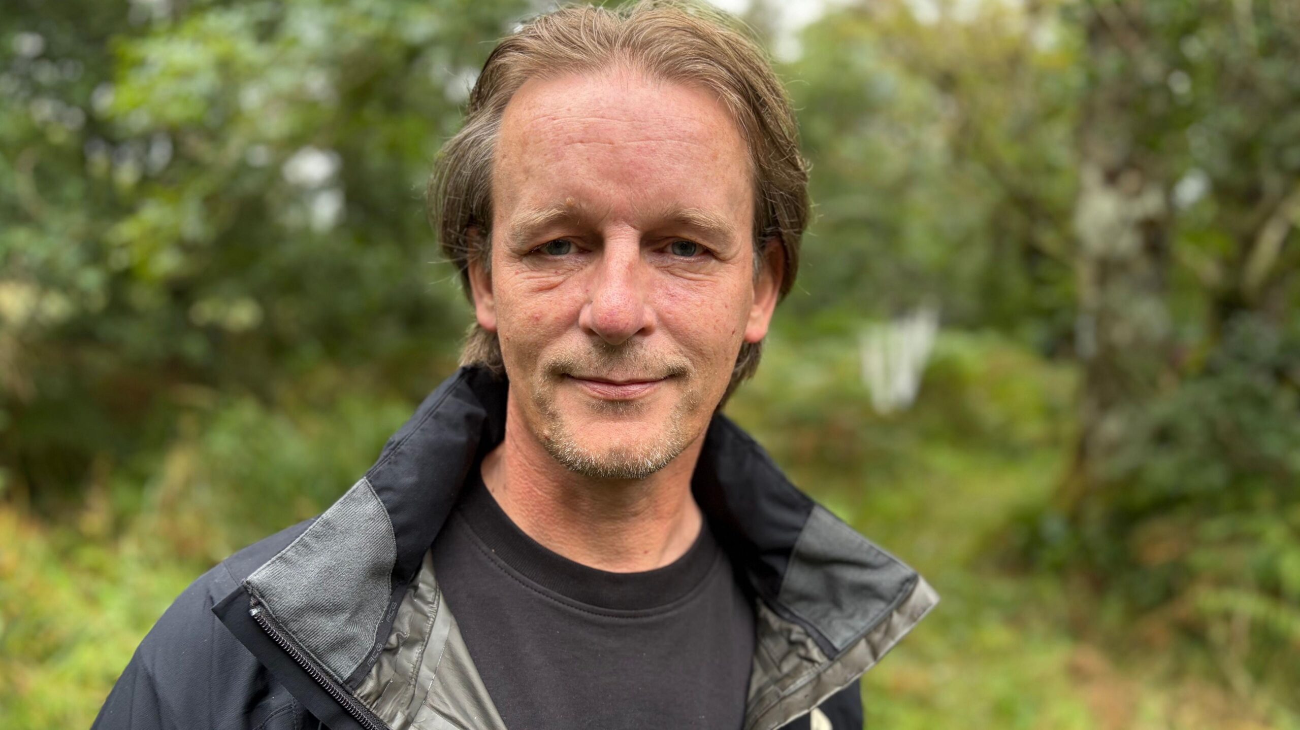 Portrait of a man wearing a dark outdoor jacket and black shirt, standing in a green wooded area with trees and foliage blurred in the background.