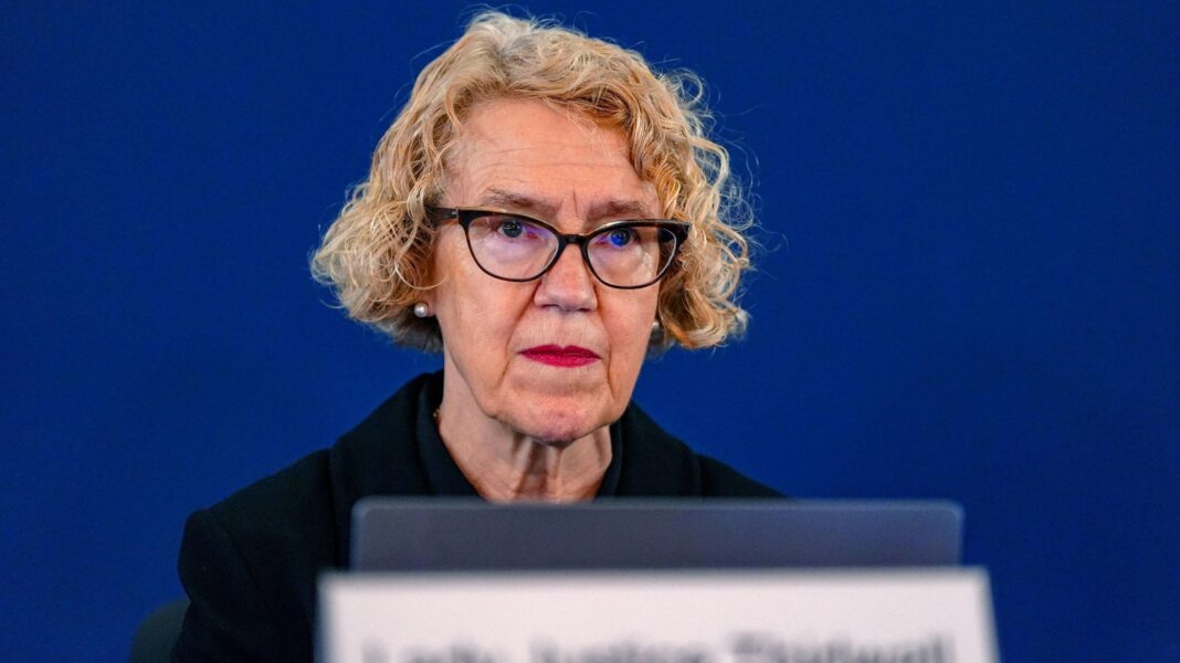 Lady Justice Kate Thirlwall, a middle-aged woman with short blonde curly hair and black-rimmed glasses, sits behind a laptop at Liverpool town hall. The background is dark blue.