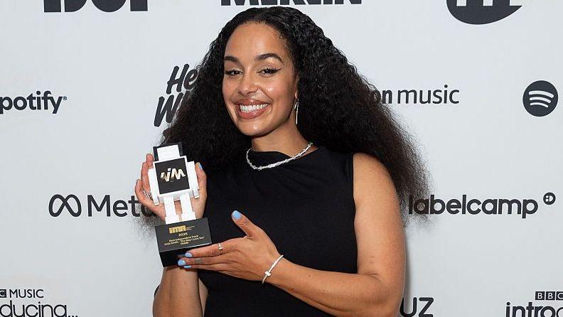 Jorja Smith wears a sleeveless black dress paired with a silver necklace and bracelet. Her nails are painted blue and she holds her award while smiling at the camera. She has long thick black hair which is parted in the middle and sits past her shoulders