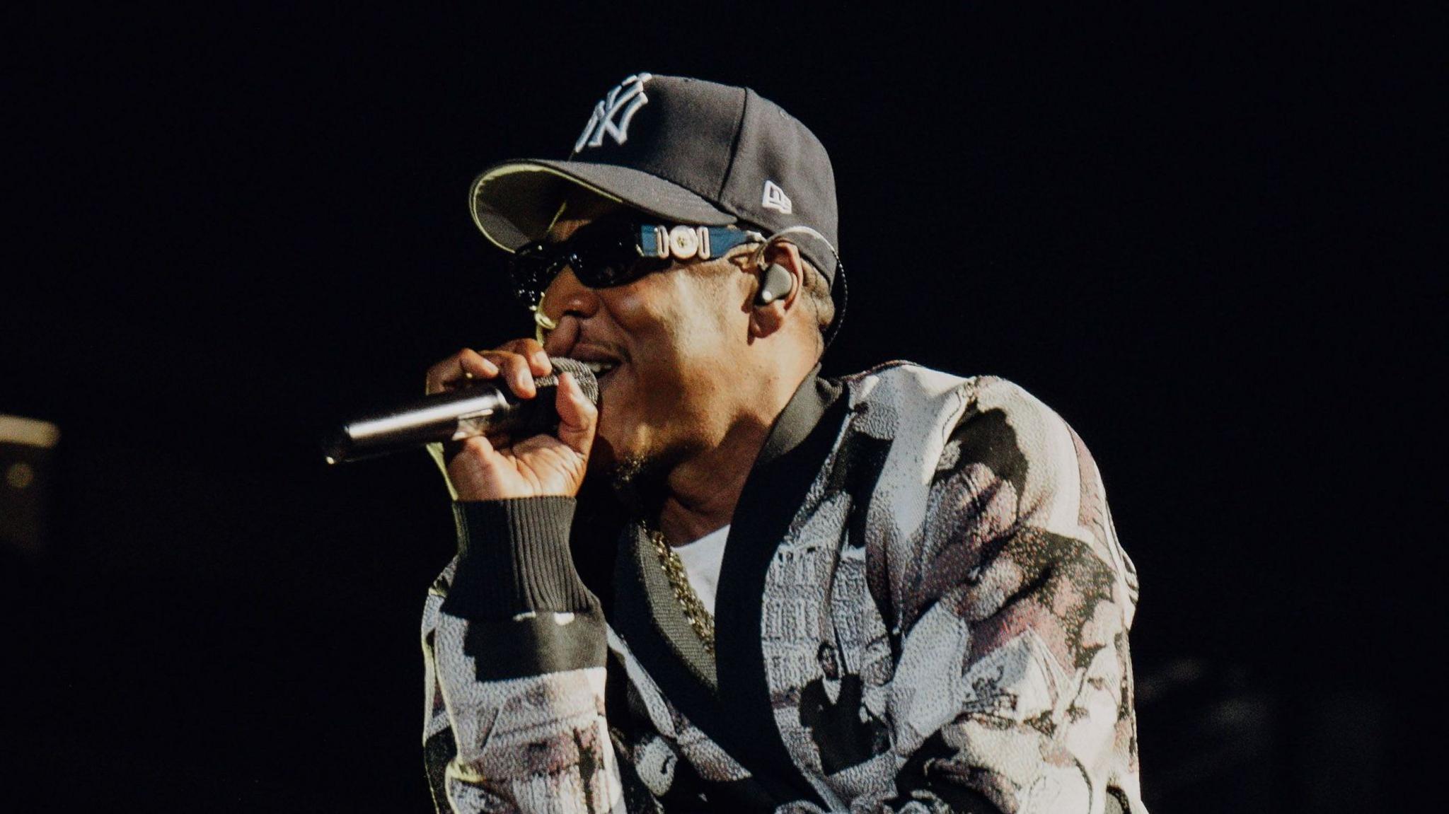 Jay-Z raps into a microphone while performing on stage. He wears a black and white cardigan and a New York Yankees hat.