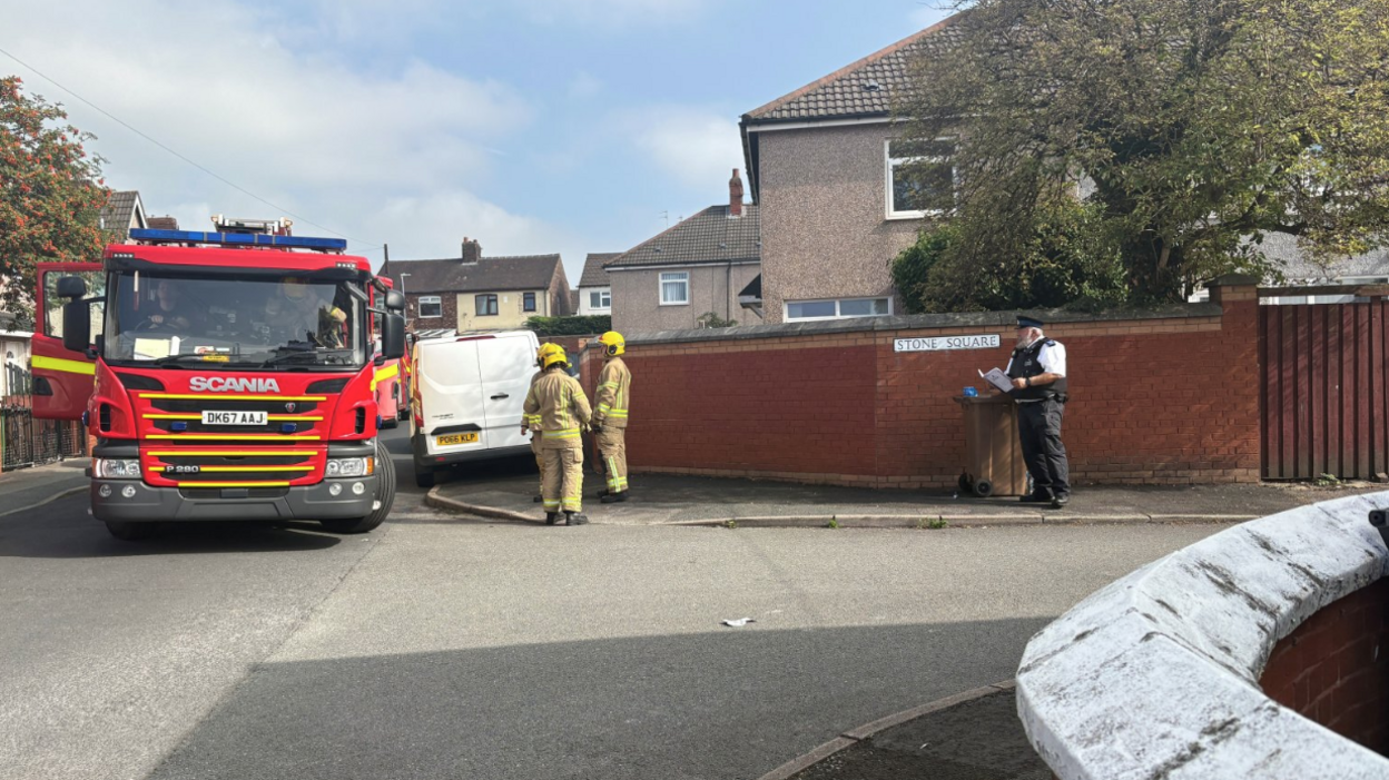 A fire engine is parked on a road outside a row of houses. A police officer and two firefighters are standing close by.