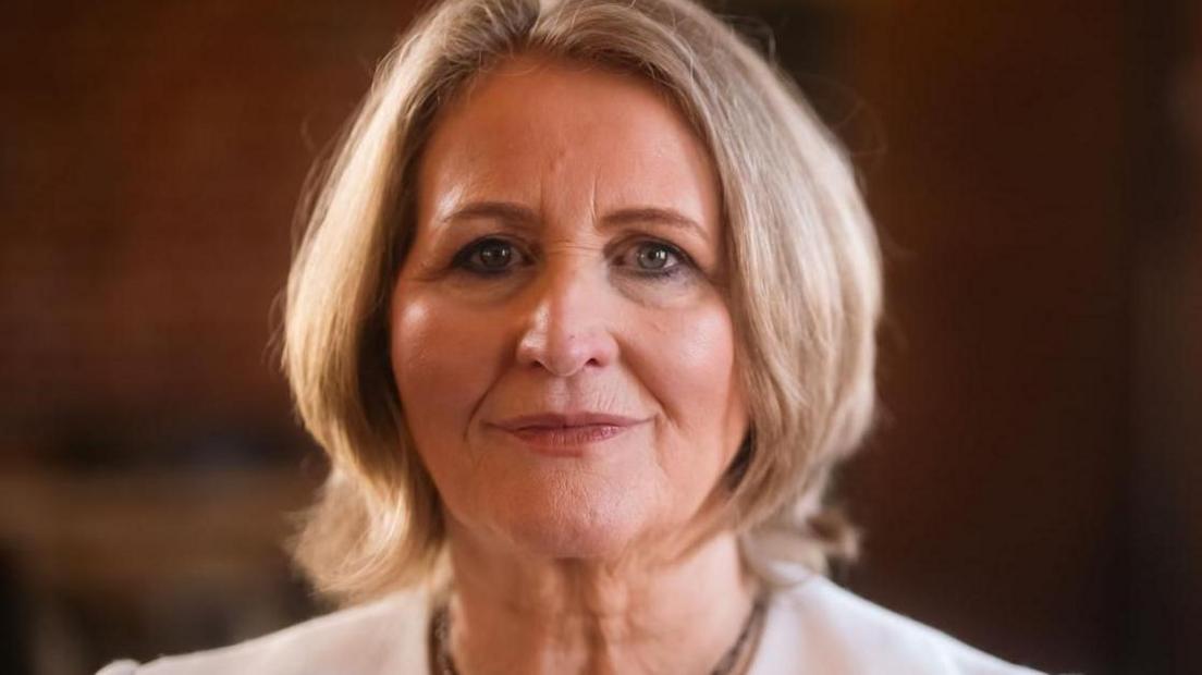 A woman with short blonde hair wearing a white suit jacket smiles faintly at the camera