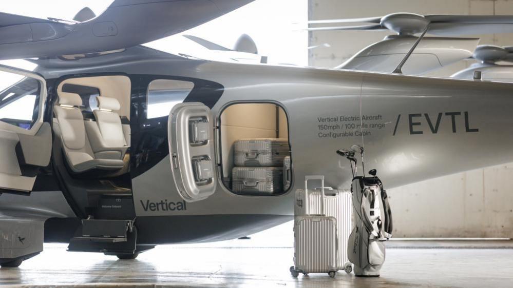 The Valo air taxi in a hangar with luggage - two silver-coloured suitcases and a white golf bag - in front of it. The doors on the side of the aircraft are open showing two seats and a luggage compartment in which a few silver-coloured suitcases can be seen. The interior, including the seats, is white/cream in colour.