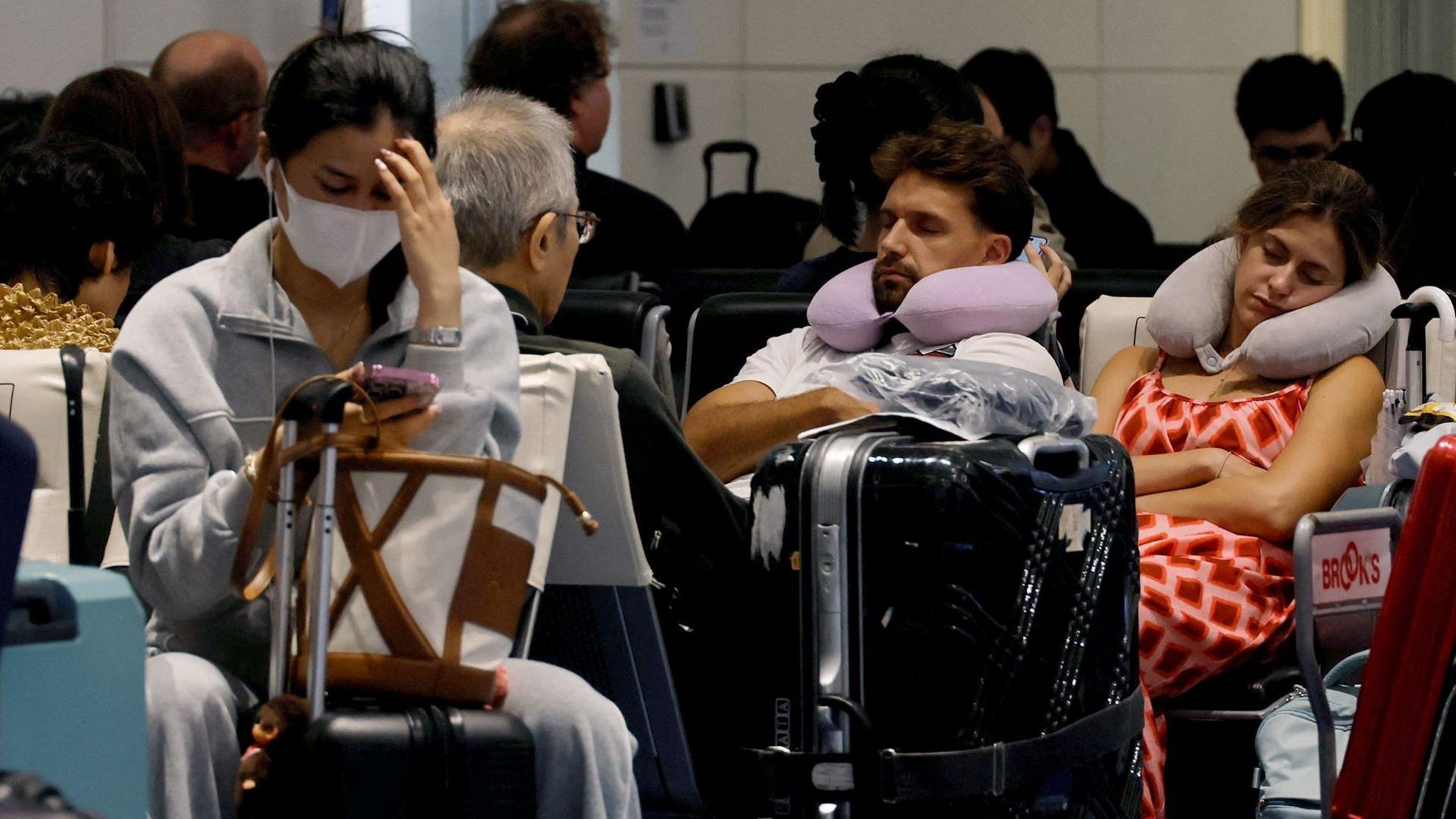 Several people sat among bags in an airport departure lounge. A woman with a medical face mask on looks down at a smart phone, a man and woman are asleep behind her.