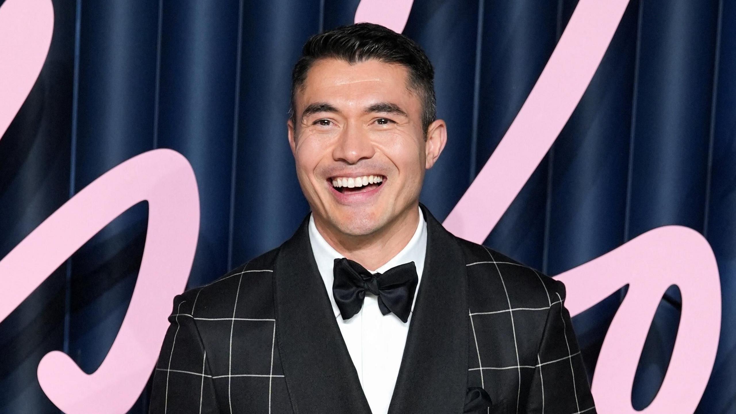 Henry Golding smiling at a premiere event, wearing a bow tie and black jacket.