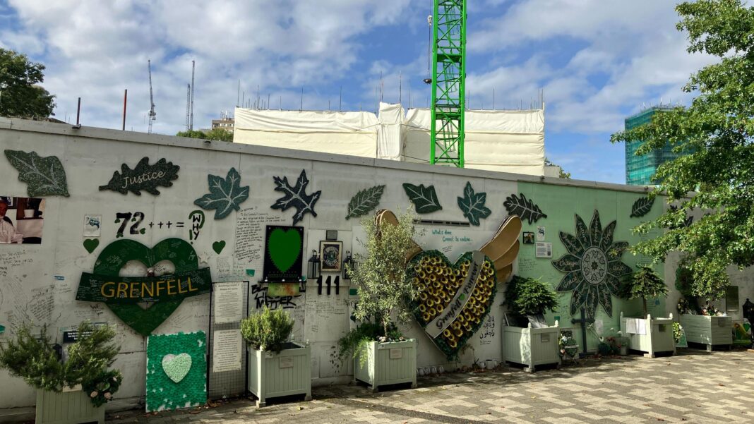A Grenfell memorial wall covered with green hearts, tributes and the number 72, with plants and flowers in front and construction work visible behind.