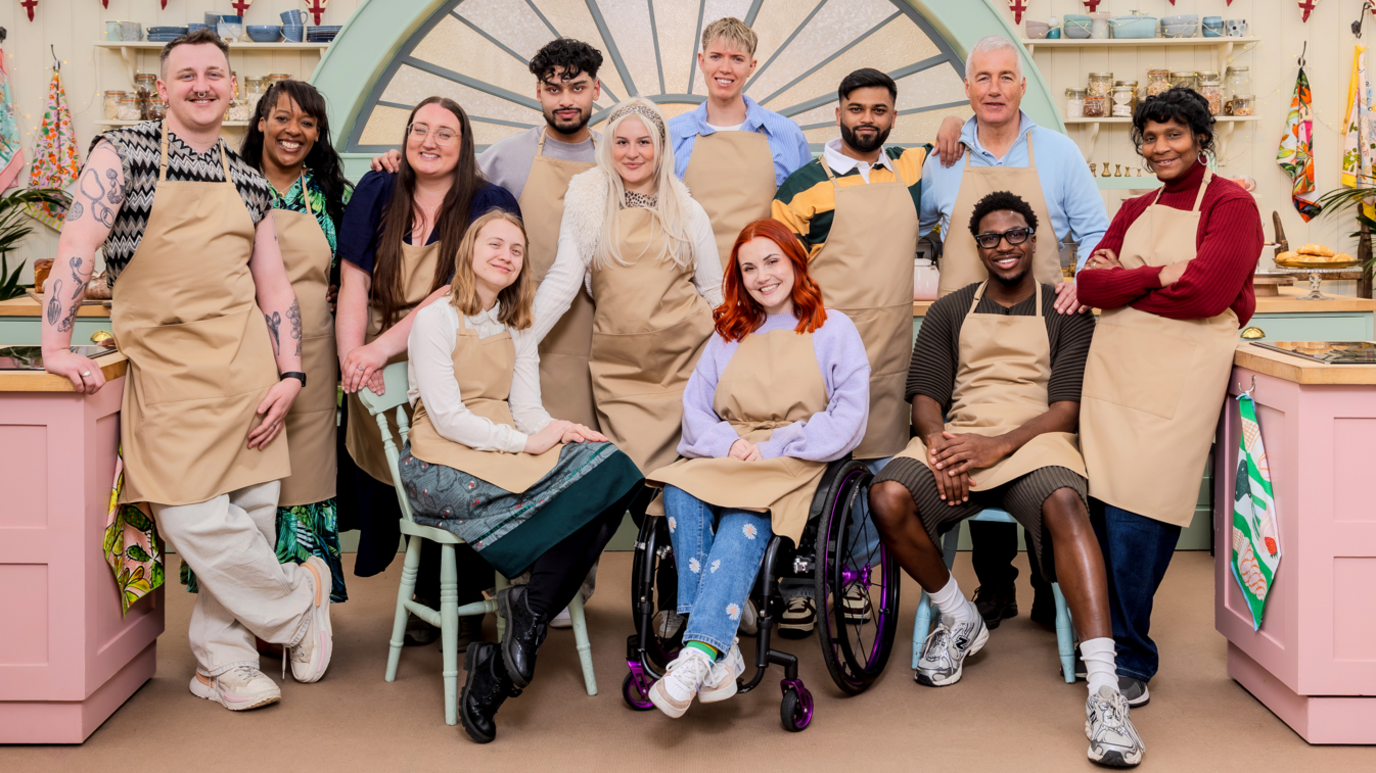 Twelve contestants pose in the pink Bake Off kitchen