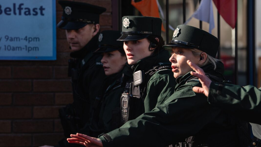 Shane, Aisling, Annie and Grace are all standing in a line in dark green police uniforms. They have hats and stab vests on with walkie-talkies. Grace has her hand out alongside another officer who is mostly out of shot. They have a shocked look on their face. They are standing in front of a gate with colourful bunting on it and next to a wall with a sign with opening times on it.