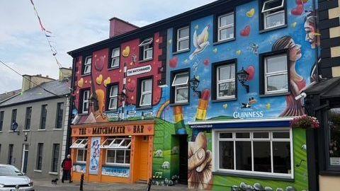 Lisdoonvarna's colourful Matchmaker Bar stands out from the typical Irish street, its exterior painted in orange, blue and other shades for a mural showing sheep, the Guinness mascot, a rainbow and other images. A man walks his dog in front of the pub while a car passes