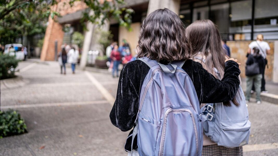 Two university students with purple backpacks walk across a university campus. Only the backs of their heads are visible. One has short wavy dark hair and wears a black velvet jacket. She has her right arm around the second student, who has long dark hair.
