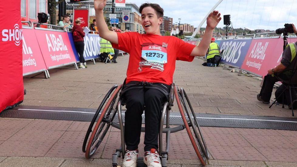 A young person sitting in a wheelchair with arms raised, and wearing a red t-shirt with a race number. Behind can be seen part of the route for the "AJ Bell Great North Run junior & mini", with barriers to either side, lined by posters.