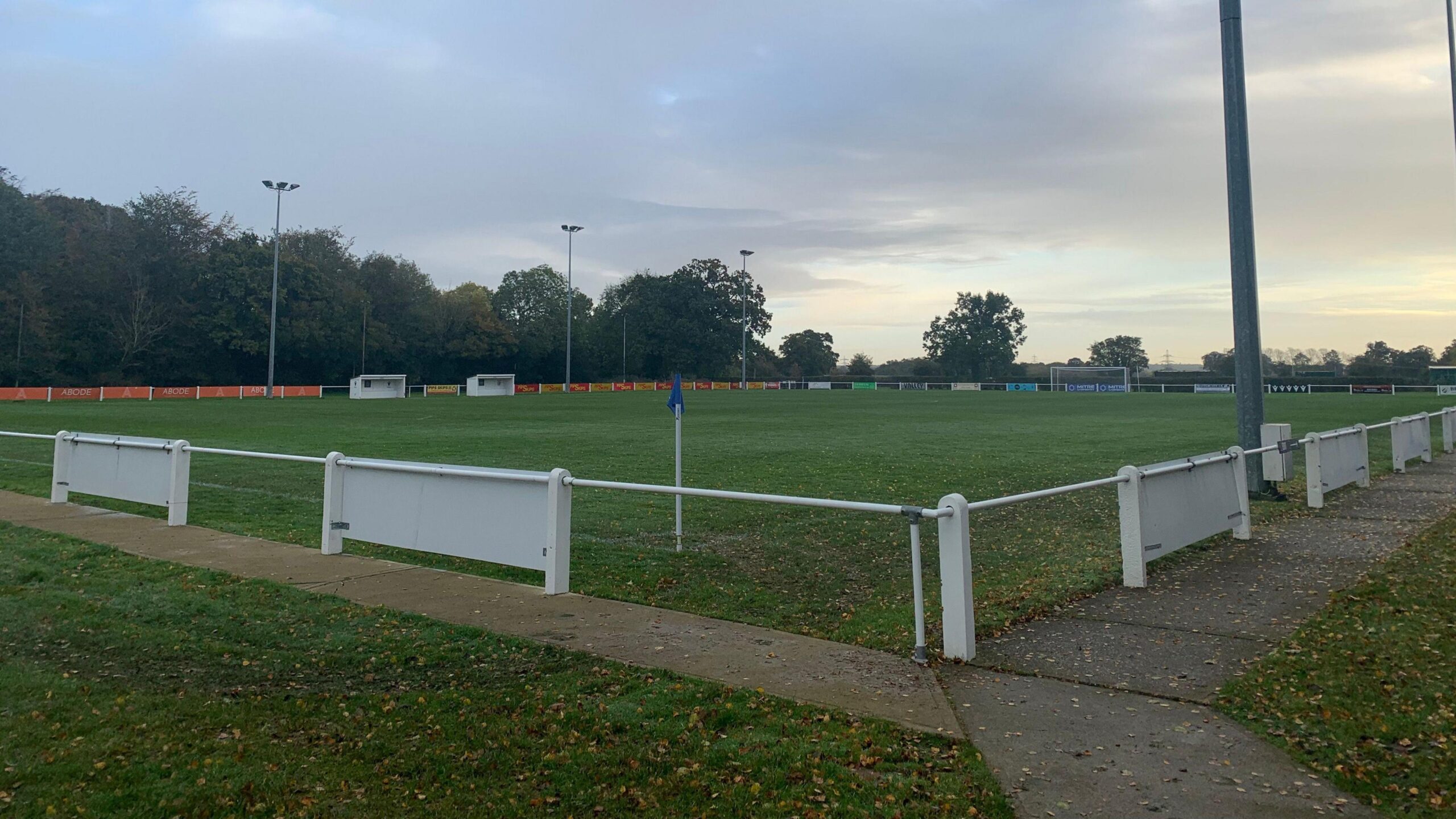 Football pitch with low, bar fence and boards around the ground. Two football dugouts are at the side of the pitch. A goal is at one end. Trees surround the pitch.