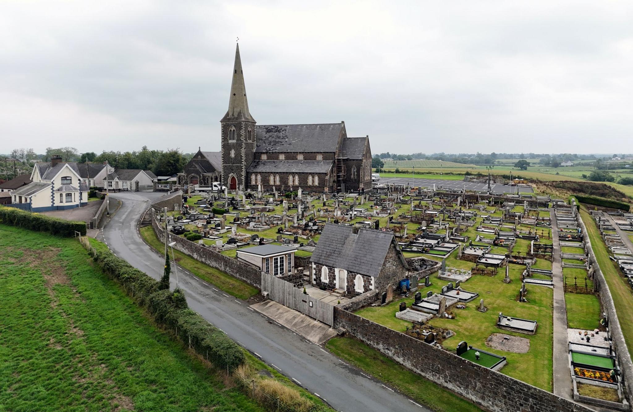 An aerial shot of a large church with a spire, alongside numerous graves on a countryside plot