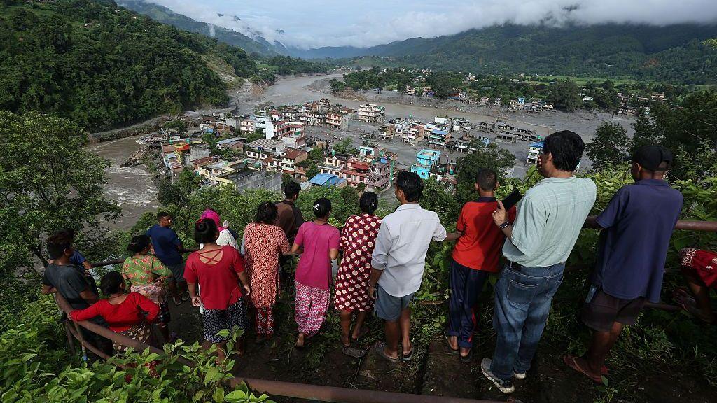 Residents stand on a hillside lookout overlooking a town heavily damaged by flooding. A muddy river cuts through the valley, with debris and sediment covering large parts of the settlement and surrounding riverbanks. Forested mountains rise on both sides under low clouds, while people gather along a railing in the foreground to view the destruction below.