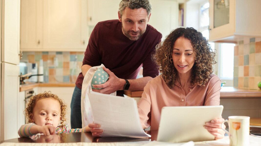 Almost half of households do not see benefits of economic growth, report says A family of three looking at bills and finances at a kitchen table. From left to right, a child is standing on the floor looking out across the table, a man is looking over a woman's shoulder at A4 sheets of paper she is holding while standing drying a mug, and a woman sits at the table holding the papers and reading. The kitchen behind them in soft focus is mostly pastel-coloured.