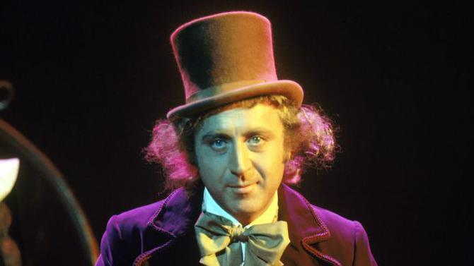 A close up of the actor Gene Wilder in character as Willy Wonka. He is wearing a large top hat and a bow tie and white shirt, and purple jacket.