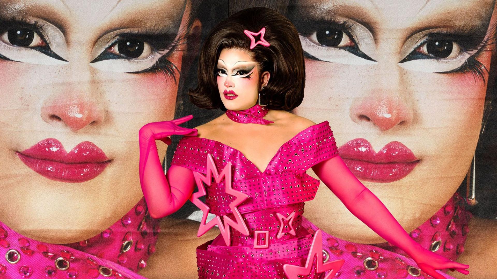 Mocha wears a fushia pink off-the-shoulder dress with matching pink gloves and has a large brunette wig on. She has dramatic eye makeup. She is standing in front of a poster board which has her face on it.