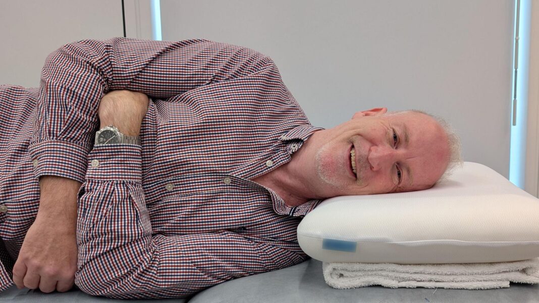 ‘I take my pillow everywhere’: An expert’s guide to finding your perfect match Person lying on their side on a padded treatment table with their head resting on a foam pillow. The person is wearing a checked button-up shirt and a wristwatch, with arms folded across the torso. The pillow is positioned on a folded towel. The window blinds in the background are drawn.