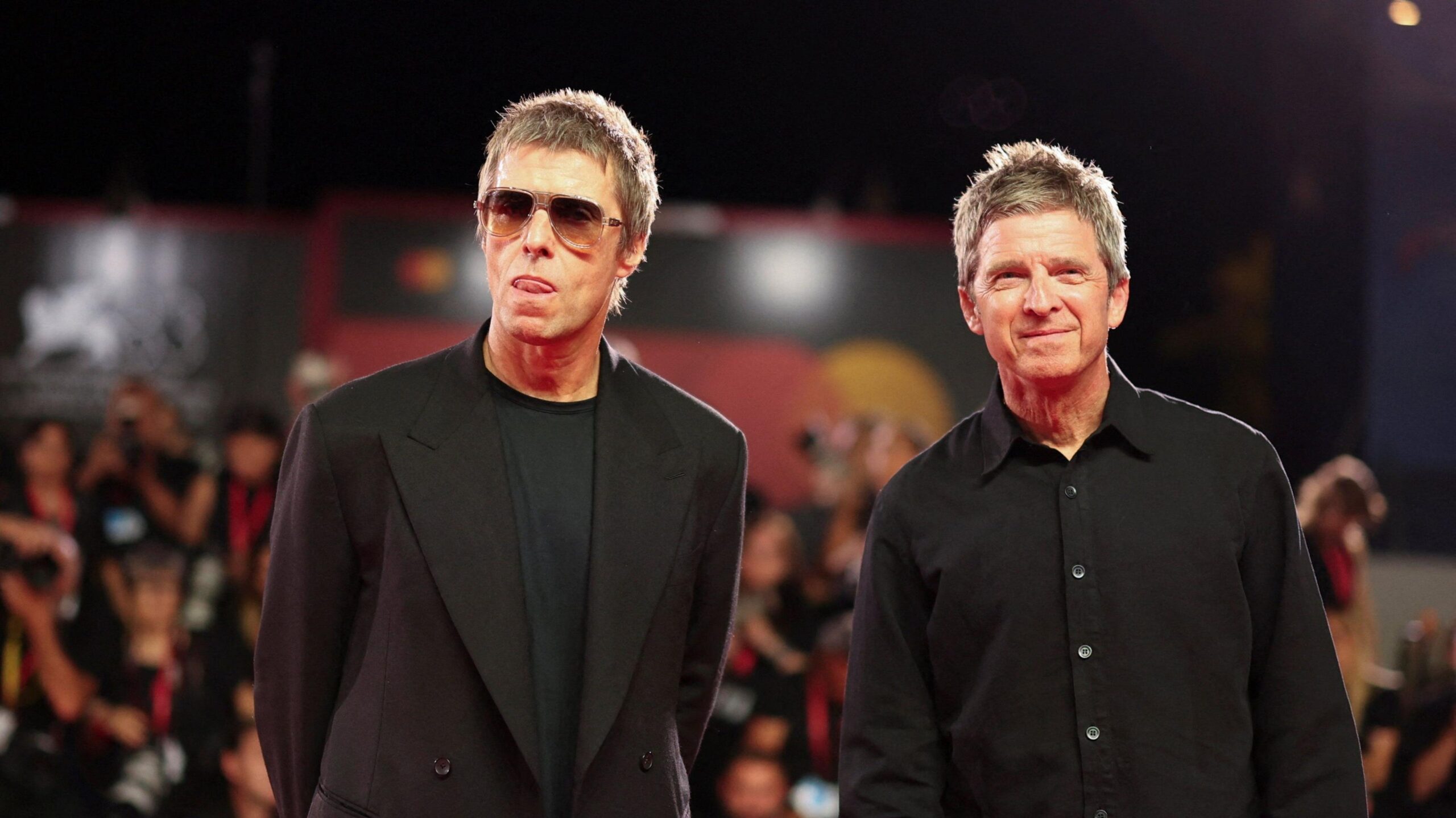 Liam and Noel Gallagher wearing black clothing stand on a red carpet with photographers stood behind them.