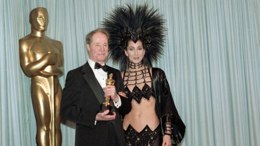 Legendary costume designer Bob Mackie dies aged 87 Cher's outfit is a black bejewelled skirt and top that shows all of her stomach, and she has a huge black headpiece on that has many black feathers sticking into the air