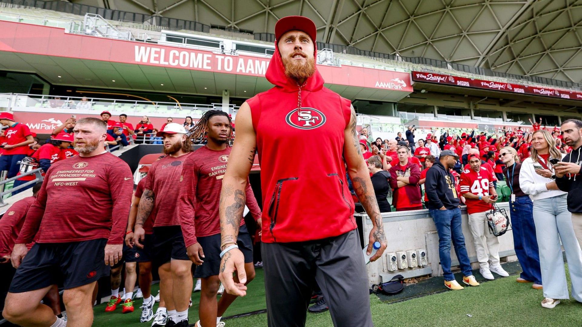 Men in red sports gear walk out of a tunnel into a stadium with fans behind them